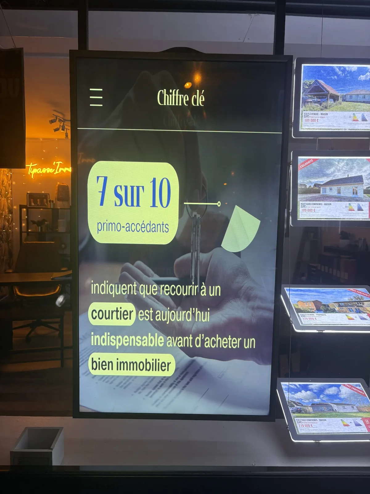 Écran LCD en vitrine d'une agence immobilière diffusant une infographie sur le recours à un courtier